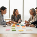 Équipe mixte de collaborateurs réunie autour d’une table, organisant des post-it colorés vierges et des éléments neutres sur un tableau de travail dans un atelier lumineux.